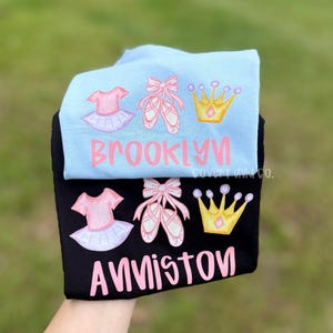 Puede incluir: Una camiseta azul claro y una camiseta negra, cada una con un gráfico de un tutú rosa, zapatillas de ballet rosas con cintas y una corona dorada. La camiseta azul claro tiene el nombre BROOKLYN en rosa y la negra tiene ANNISTON en rosa.