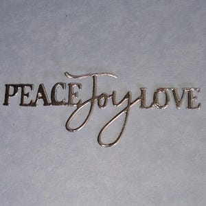 May include: Silver metal word art spelling out "PEACE Joy LOVE" in a cursive and block font style. The words are placed on a light grey background. The word "Joy" is in a cursive font, while "PEACE" and "LOVE" are in a block font.