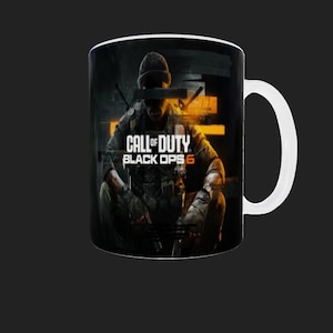 May include: A black ceramic coffee mug with a white handle. The mug features a graphic of a soldier from the Call of Duty: Black Ops 6 video game. The soldier is wearing camouflage and holding a gun. The text "Call of Duty: Black Ops 6" is printed in orange on the mug.