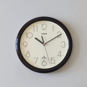 Op de afbeelding: Een ronde wandklok met een glanzend zwarte rand en een gebroken witte wijzerplaat. De klok heeft zwarte Arabische cijfers, dunne zwarte uur- en minuutwijzers en een dunne secondewijzer. CITIZEN staat onder de 12 en QUARTZ staat boven de 6.