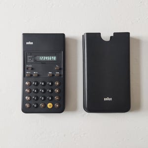 Könnte beinhalten: Ein schwarzer Braun-Taschenrechner mit der Anzeige 12345678, neben der passenden schwarzen Hülle. Der Rechner hat braune und gelbe Tasten. Der Markenname "Braun" ist auf beiden Artikeln sichtbar.