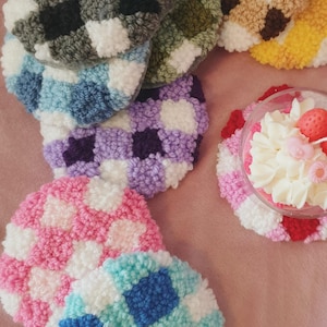 Può includere: Una collezione di sottobicchieri rotondi fatti a mano in vari colori, tra cui rosa, blu, viola e verde, con un motivo a scacchi. I sottobicchieri sono realizzati in un materiale morbido e strutturato. È visibile anche un dessert con panna montata e una fragola.