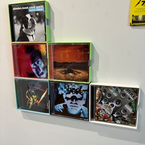 May include: A wall display of six CD cases with various album covers. The top case features a black and white photo of a dog with the text "eskAUBEi & Tomek Nowak Quartet bedzie dobrze". The other cases feature a variety of colourful images, including a red and blue image of a person, a red and orange image of a desert landscape, a black and white image of a person, a black and white image of a dog, and a colourful image of a DJ with speakers.