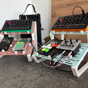 Puede incluir: Tres soportes blancos en forma de escalón con plataformas de diferentes colores. El soporte superior tiene un sintetizador negro y naranja, el soporte del medio tiene un sintetizador negro y verde, y el soporte inferior tiene una calculadora negra y una calculadora transparente. Todos los soportes tienen clips de plástico blanco.