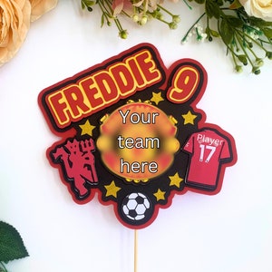 Puede incluir: Un adorno para tarta con temática de fútbol en rojo y negro con el nombre "FREDDIE" y el número "9". El diseño incluye un balón de fútbol, una camiseta con el número 17 y un espacio para "Your team here".