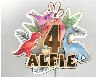May include: A gold number four cake topper with a red dinosaur, a blue dinosaur, and a purple flying dinosaur. The topper also includes the name "Alfie" in gold letters.
