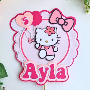 May include: Pink and white Hello Kitty cake topper with a pink bow, a pink flower, and the number 5 on a pink heart-shaped balloon. The topper also includes the name "Ayla" in pink letters.