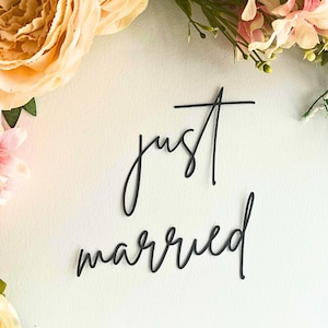 Könnte beinhalten: Schwarzer Schriftzug "just married" auf weißem Hintergrund, umgeben von pfirsichfarbenen und rosa Blumen und Grün. Ein dekoratives Element für Hochzeiten oder Feiern.