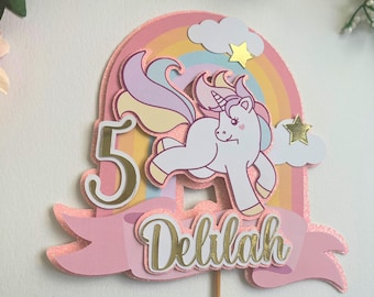 May include: A pink and gold glittery unicorn cake topper with a rainbow, clouds, and stars. The number 5 and the name "Delilah" are also featured. The unicorn has a white body, pink cheeks, and a colorful mane and tail.