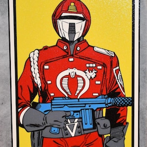 May include: A framed illustration of a character in a red uniform with a white helmet and a blue weapon, set against a yellow background. The character has a white cobra emblem on the chest. The frame is white with a black border.