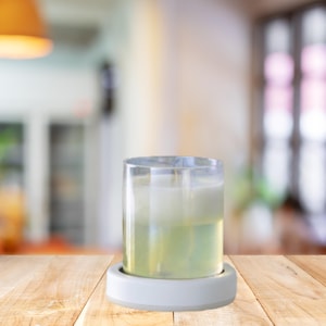 May include: A clear glass candle holder with a white base. The candle inside has a layered appearance, with a light green liquid at the bottom and a white solid wax at the top. The holder sits on a wooden surface.