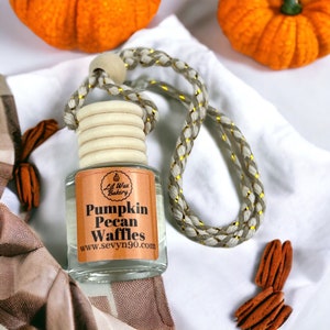 May include: A clear glass air freshener bottle with a wooden cap and a braided cord. The label reads "Pumpkin Pecan Waffles" and "Lil Wax Bakery". The background includes orange pumpkins and pecans.
