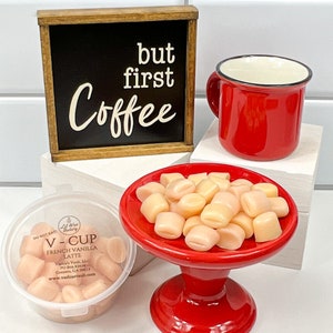 Op de afbeelding: Een rode mok met een zwarte rand, een klein rood voetstuk met een witte keramische schaal gevuld met lichtroze marshmallows en een houten bord met de tekst "but first coffee" in witte letters op een zwarte achtergrond.