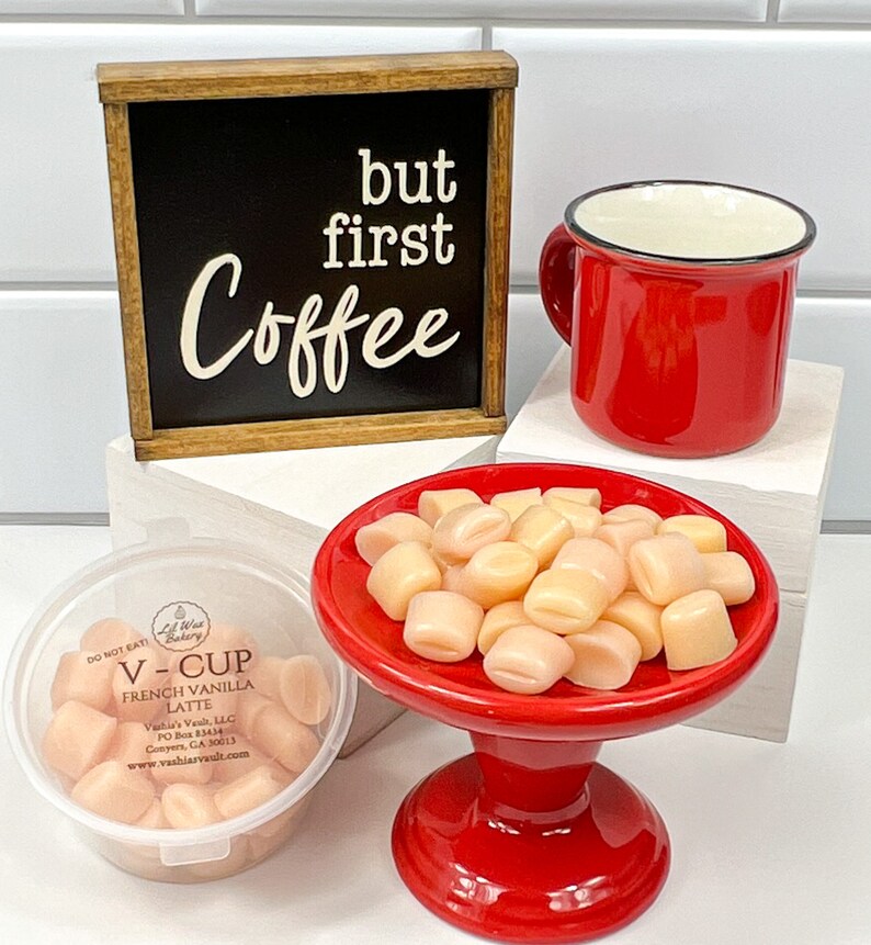 Op de afbeelding: Een rode mok met een zwarte rand, een klein rood voetstuk met een witte keramische schaal gevuld met lichtroze marshmallows en een houten bord met de tekst "but first coffee" in witte letters op een zwarte achtergrond.