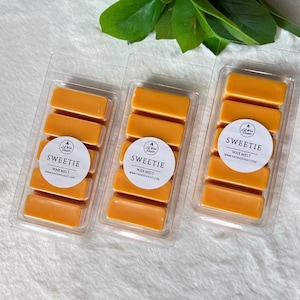 May include: Three clear plastic containers with orange wax melts. Each container has a white circle label with the text "Sweetie Wax Melt" and a website address.
