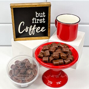 Op de afbeelding: Een rode koffiemok met een zwarte rand, een klein rood voetstuk met een stapel chocolade snoepjes en een houten bord met de tekst "but first coffee" op een witte betegelde achtergrond.
