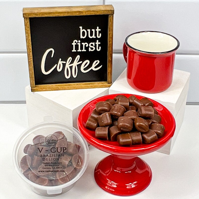 Op de afbeelding: Een rode koffiemok met een zwarte rand, een klein rood voetstuk met een stapel chocolade snoepjes en een houten bord met de tekst "but first coffee" op een witte betegelde achtergrond.