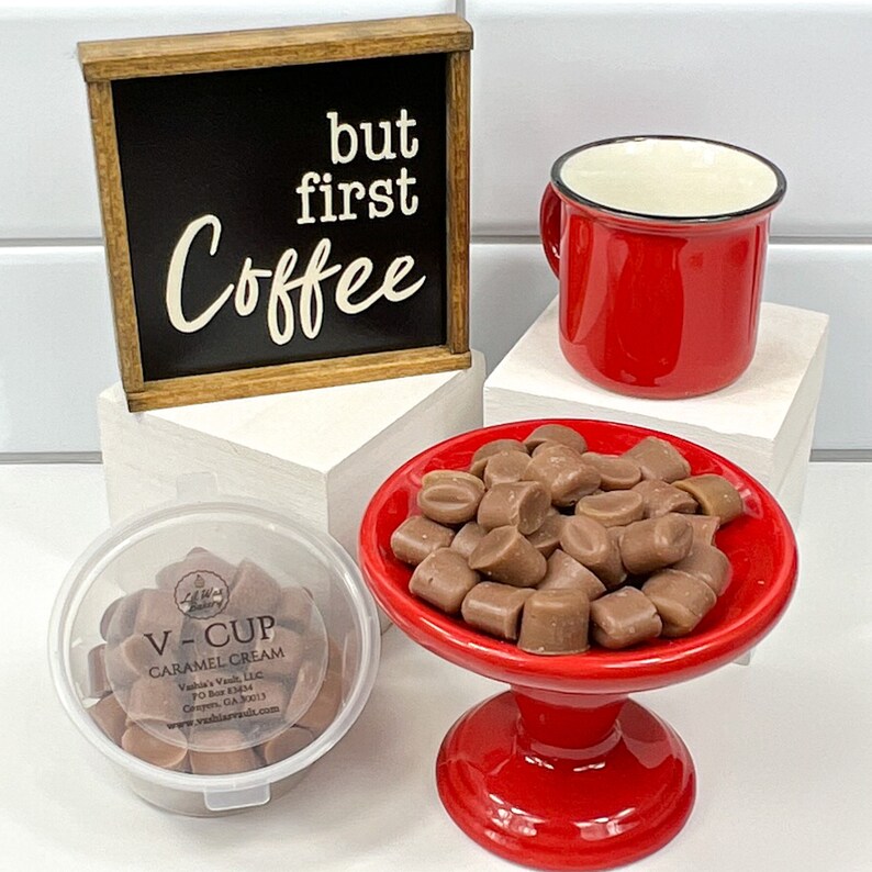 Op de afbeelding: Een rode koffiemok, een klein rood voetstuk met een stapel bruine karamel snoepjes en een doorzichtige plastic container met meer karamel snoepjes. De container heeft een label met de tekst "V-CUP CARAMEL CREAM" en een bord met de tekst "but first Coffee".