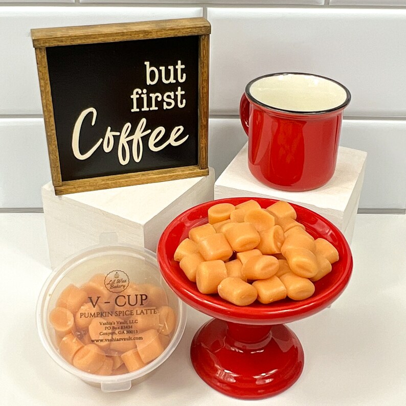 Op de afbeelding: Een rode mok met een zwarte rand en een witte binnenkant staat op een wit oppervlak naast een houten bord met de tekst "but first coffee". Een rood voetstuk houdt een kleine kom gevuld met oranje was smelt. Een doorzichtige container met een label met de tekst "V-CUP PUMPKIN SPICE LATTE" bevat meer oranje was smelt.