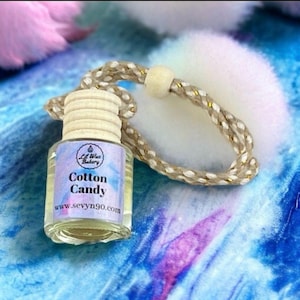 May include: A small glass bottle with a wooden cap and a gold string hanging from it. The bottle is filled with a clear liquid and has a label that says "Lil Wax Bakery Cotton Candy www.sevyn90.com".