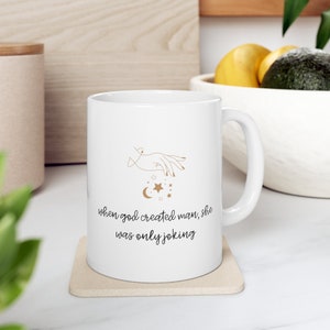 Puede incluir: Taza de cerámica blanca con un contorno dorado de una mano con estrellas y una luna creciente. El texto "when god created man, she was only joking" está impreso en la taza.
