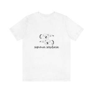 May include: White t-shirt featuring a black graphic of two hands with moon and star designs, and the text "summon serotonin" in a handwritten style. The shirt is a casual, everyday wear item.