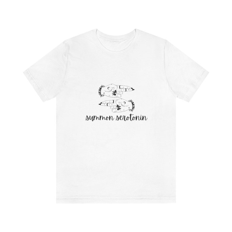 May include: White t-shirt featuring a black graphic of two hands with moon and star designs, and the text "summon serotonin" in a handwritten style. The shirt is a casual, everyday wear item.