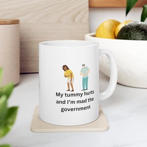 May include: White ceramic mug with a cartoon illustration of a person with a stomach ache and another person standing nearby. The mug has the text "My tummy hurts and I'm mad the government" printed on it.
