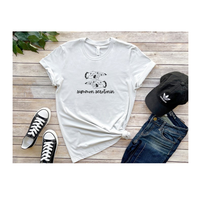 May include: A white t-shirt with the phrase "summon serotonin" printed in black. The shirt features a graphic of two hands with a floral design. A black baseball cap and blue jeans are also in the image.