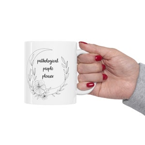 May include: White ceramic mug with a black crescent moon design and the text "pathological people pleaser" inside the moon. The mug has a black floral design around the moon.