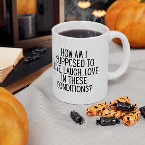 May include: White ceramic mug filled with dark liquid, featuring the text "HOW AM I SUPPOSED TO LIVE, LAUGH, LOVE IN THESE CONDITIONS?". Halloween-themed candies in orange and black are scattered around the mug. A pumpkin and candles are in the background.
