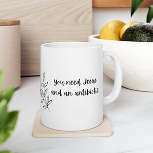 May include: White ceramic mug with a black floral design and the text "you need Jesus and an antibiotic".