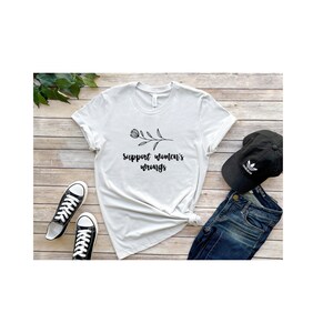 May include: A white t-shirt with a black floral design and the text "support women's wrongs". The shirt is laid out on a wooden surface with a pair of black sneakers, a pair of blue jeans, and a black baseball cap.