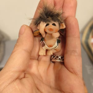Puede incluir: Una pequeña y caprichosa figura de una criatura de hadas con tez bronceada, cabello castaño y pequeñas alas. La figura lleva un pañal y tiene un anillo en el dedo. El hada se lleva la mano a la cara.