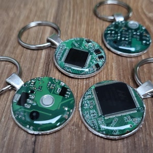 Könnte beinhalten: Schlüsselanhänger mit einem kreisförmigen Design mit grünem Leiterplattenmuster. Jeder Schlüsselanhänger hat einen silberfarbenen Metallring und eine kleine, silberfarbene Metallschlaufe. Das Leiterplattendesign umfasst schwarze rechteckige Komponenten.