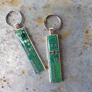 Könnte beinhalten: Zwei silberne Schlüsselanhänger mit grünen Platinen-Designs. Die Schlüsselanhänger sind rechteckig und haben oben einen silbernen Ring.