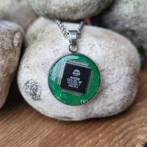 Pode incluir: Um colar de corrente prateada com um pingente redondo que apresenta um design de placa de circuito verde. A placa de circuito tem um chip preto com o texto "AUG6368 D33-JEL-NP H1SS39-1 0651ES".