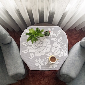 May include: A white crocheted table runner with a floral design, a small potted succulent, and a cup of coffee on a wooden table. Two grey armchairs are on either side of the table.