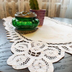 May include: A white oval lace tablecloth with a delicate floral design. The tablecloth has a scalloped edge and is decorated with intricate lacework. A green glass jar with a silver lid sits on top of the tablecloth.
