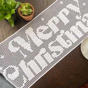 Op de afbeelding: Een witte gehaakte tafelloper met de woorden "Gather Together" in een decoratief lettertype.