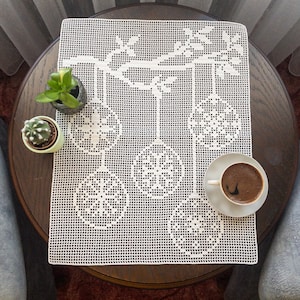 May include: A white, crocheted table runner with a Christmas ornament design, on a dark wooden table. The runner features a branch with hanging ornaments. A small potted plant and a cup of coffee are also on the table.