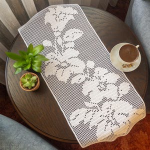 Peut inclure: Un chemin de table en crochet blanc avec un motif floral. Le chemin de table est posé sur une table en bois brun foncé avec une tasse de café et deux petits pots de plantes.
