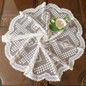 May include: White crocheted table runner with a delicate floral pattern. The runner is laid out on a wooden table with a small white teacup and saucer on a green leaf-shaped plate.
