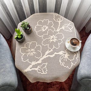 May include: A cream-coloured, octagonal lace tablecloth with a floral design on a small, round table. Two small potted plants and a cup of coffee on a saucer are on the table. Two grey armchairs surround the table.