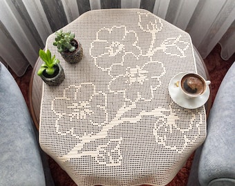 May include: An octagonal, cream-colored lace tablecloth with a floral design covers a small, round table. Two small potted plants and a cup of coffee on a saucer are on the table. The table is surrounded by two gray armchairs.