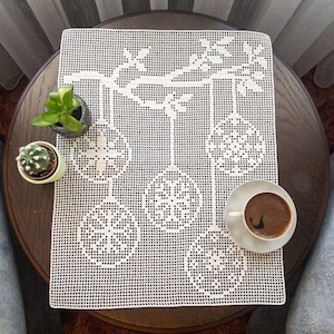 May include: A white, crocheted table runner with a detailed floral and ornament design. The runner is placed on a dark brown wooden table, with small potted plants and a cup of coffee. The design features hanging ornaments.