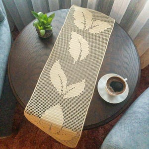 May include: A beige crocheted table runner with a leaf design on a brown wooden table. A white saucer with a cup of coffee is on the table.