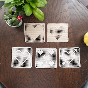 May include: Five crocheted heart-shaped coasters in a beige color. The coasters are arranged in a grid pattern on a wooden surface.