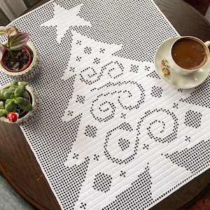 May include: White crocheted table runner with a Christmas tree design. The tree is made of circles and stars. The runner is on a wooden table with a cup of coffee and a saucer.