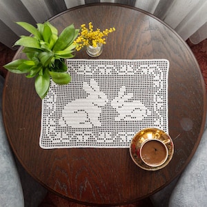 Puede incluir: Un mantel de encaje blanco con un diseño floral se encuentra sobre una mesa de madera oscura con una taza de café y un pequeño ramo de flores amarillas en un jarrón de vidrio.